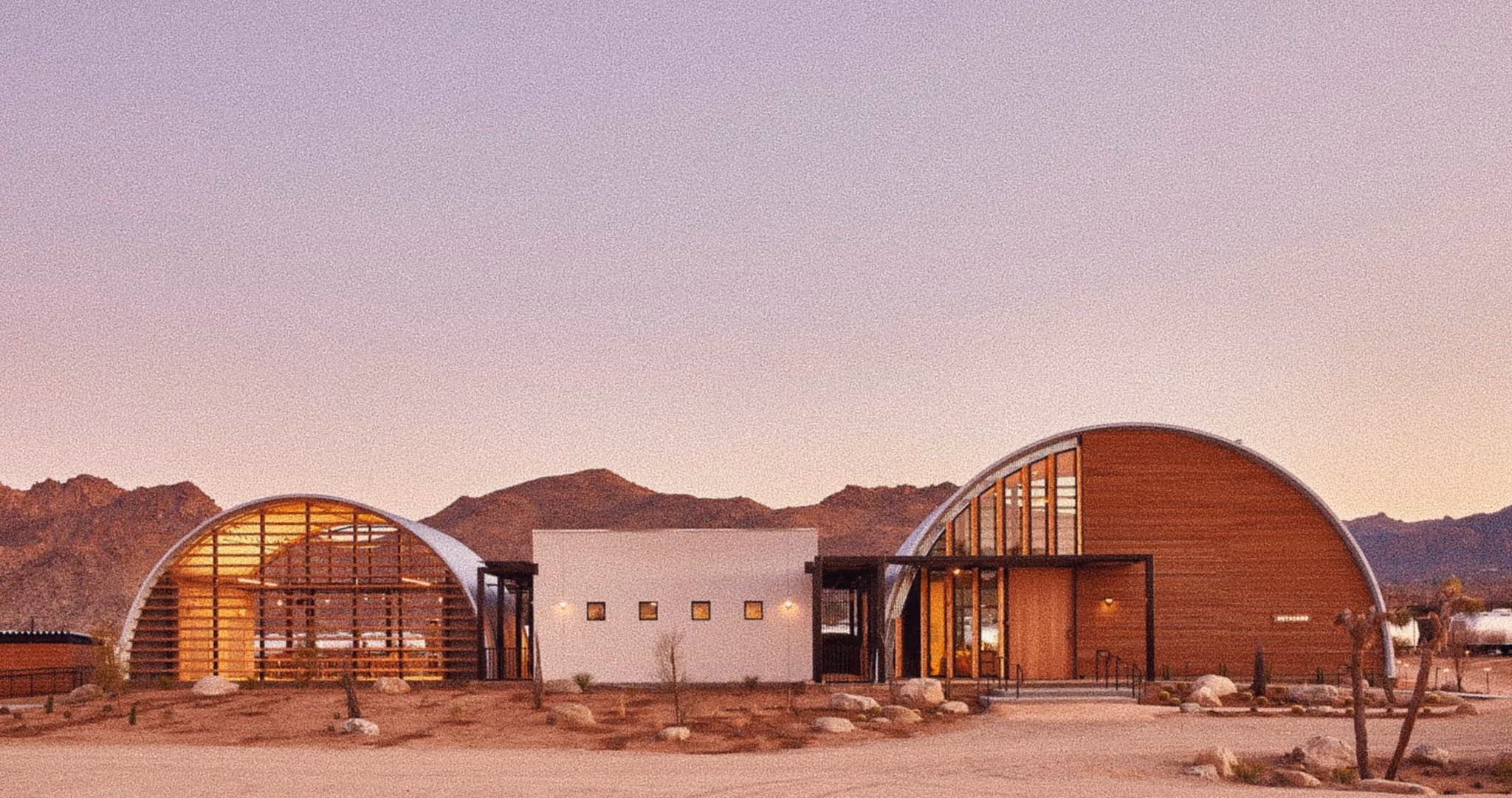 Modern building with curved roofs and wood panels set against desert mountains at dusk.