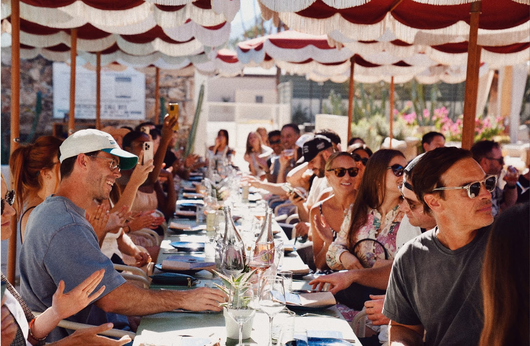 People sitting at a long outdoor table under red and white canopy enjoying a social gathering.