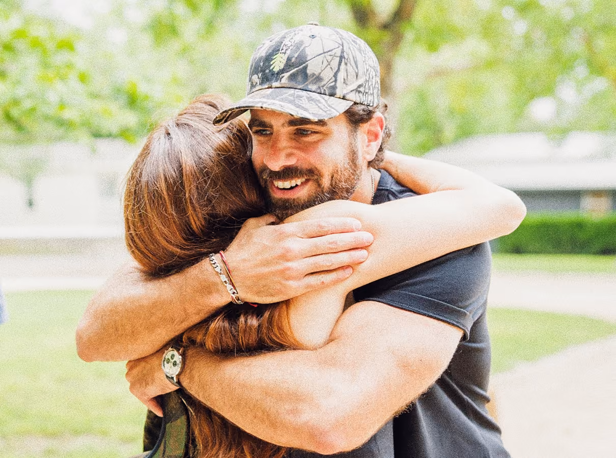 Bearded man in a cap warmly hugging a woman outdoors with greenery in the background.