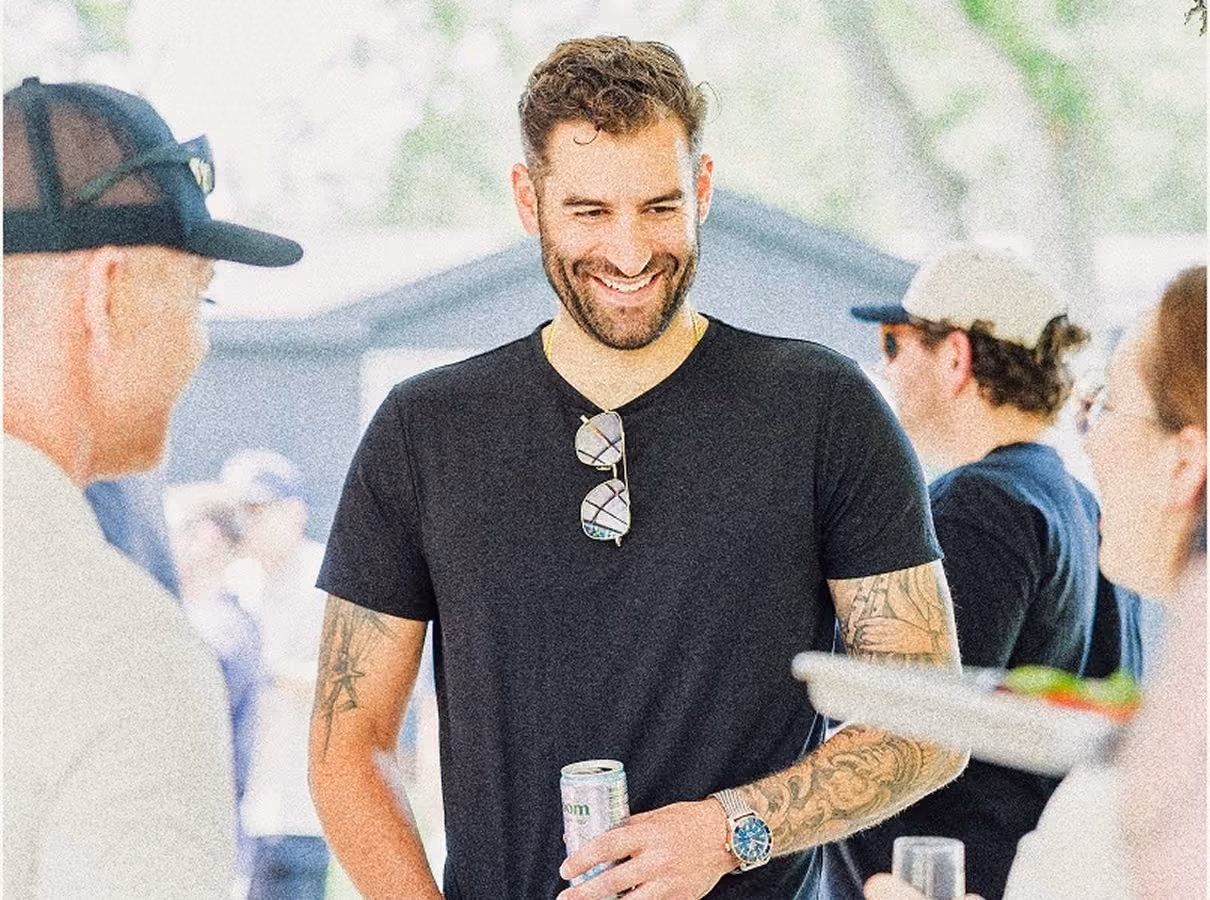 Smiling man with tattoos and glasses on his shirt holding a drink and talking to people outdoors.