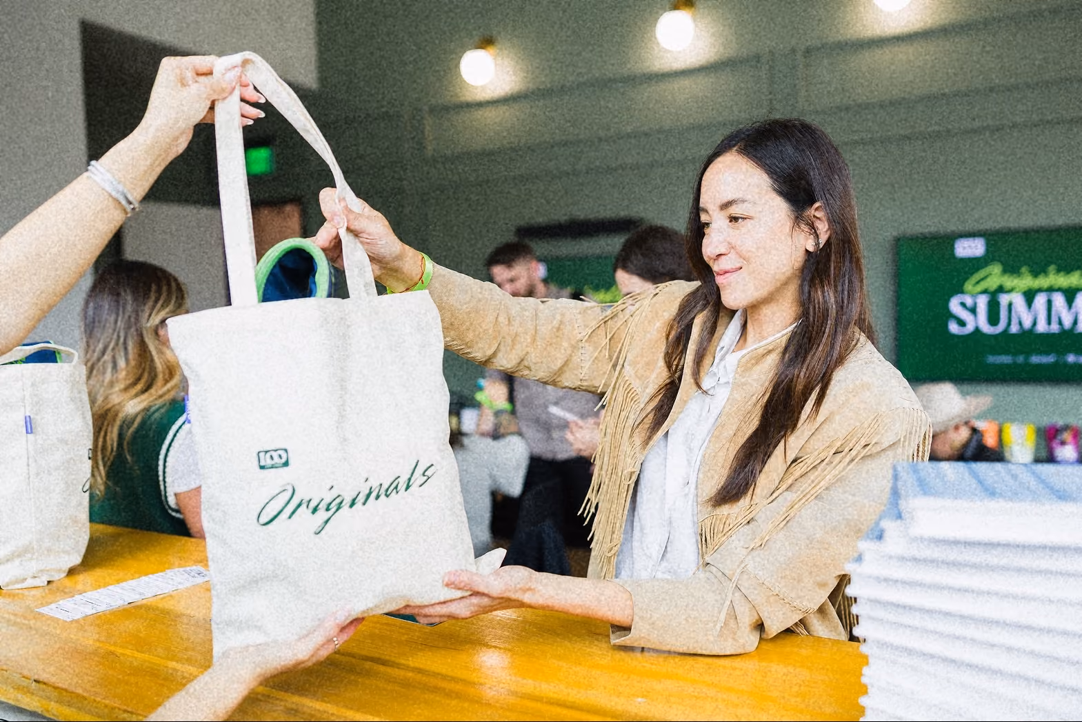 Woman handing over a tote bag labeled Originals at a summit registration desk.