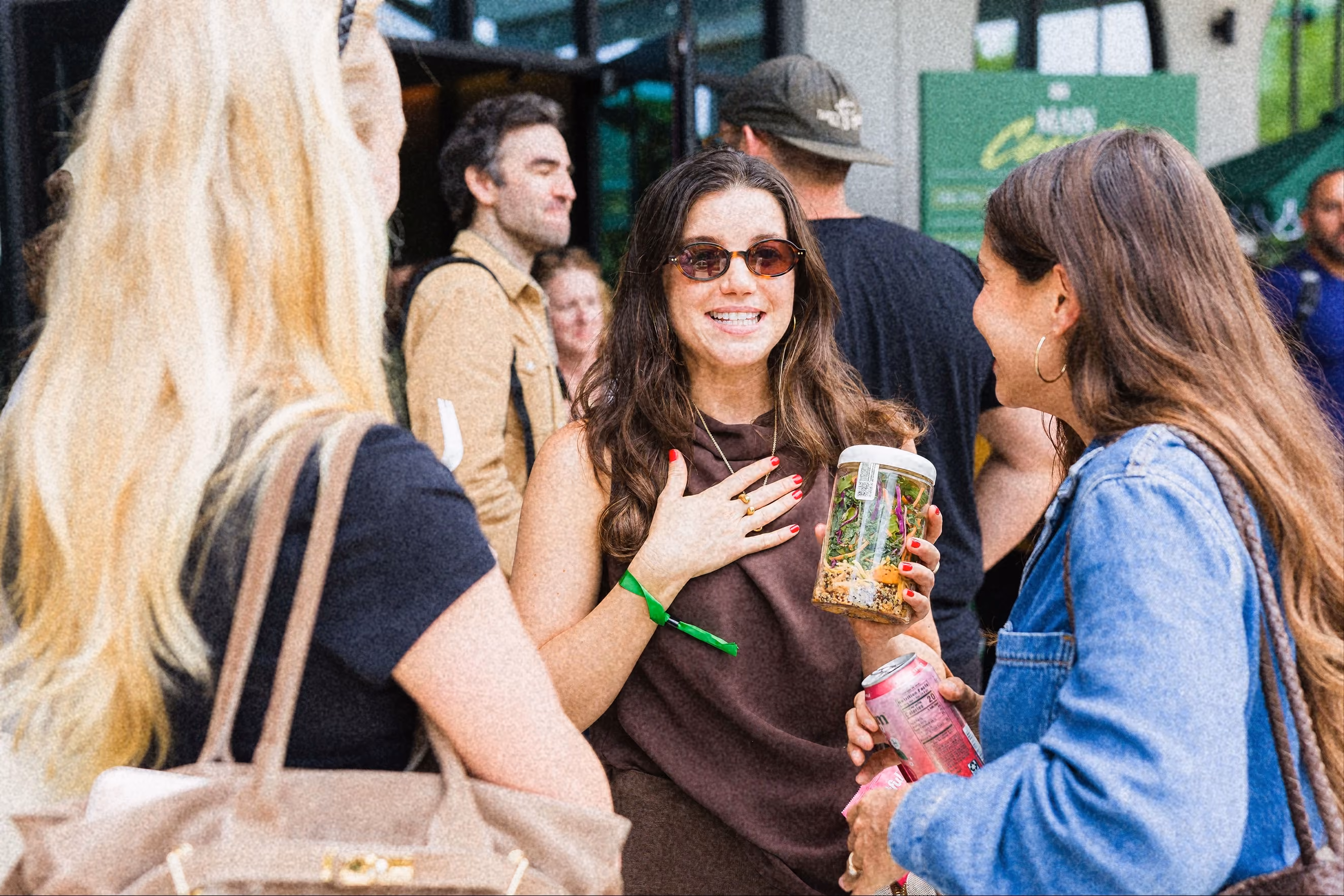 Three women engaged in conversation at an outdoor event, one holding a salad jar and another a pink drink can.