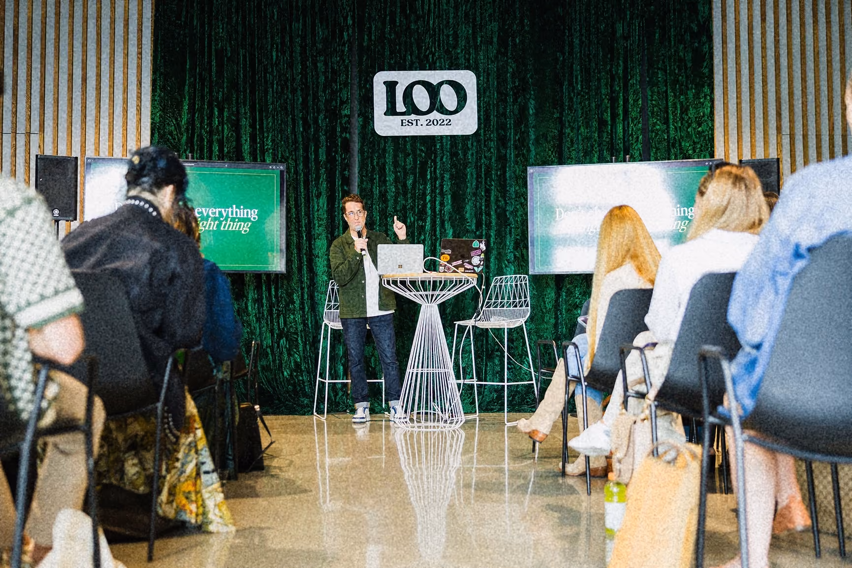 Speaker in green jacket talks at event with audience and L100 EST. 2022 sign behind him.