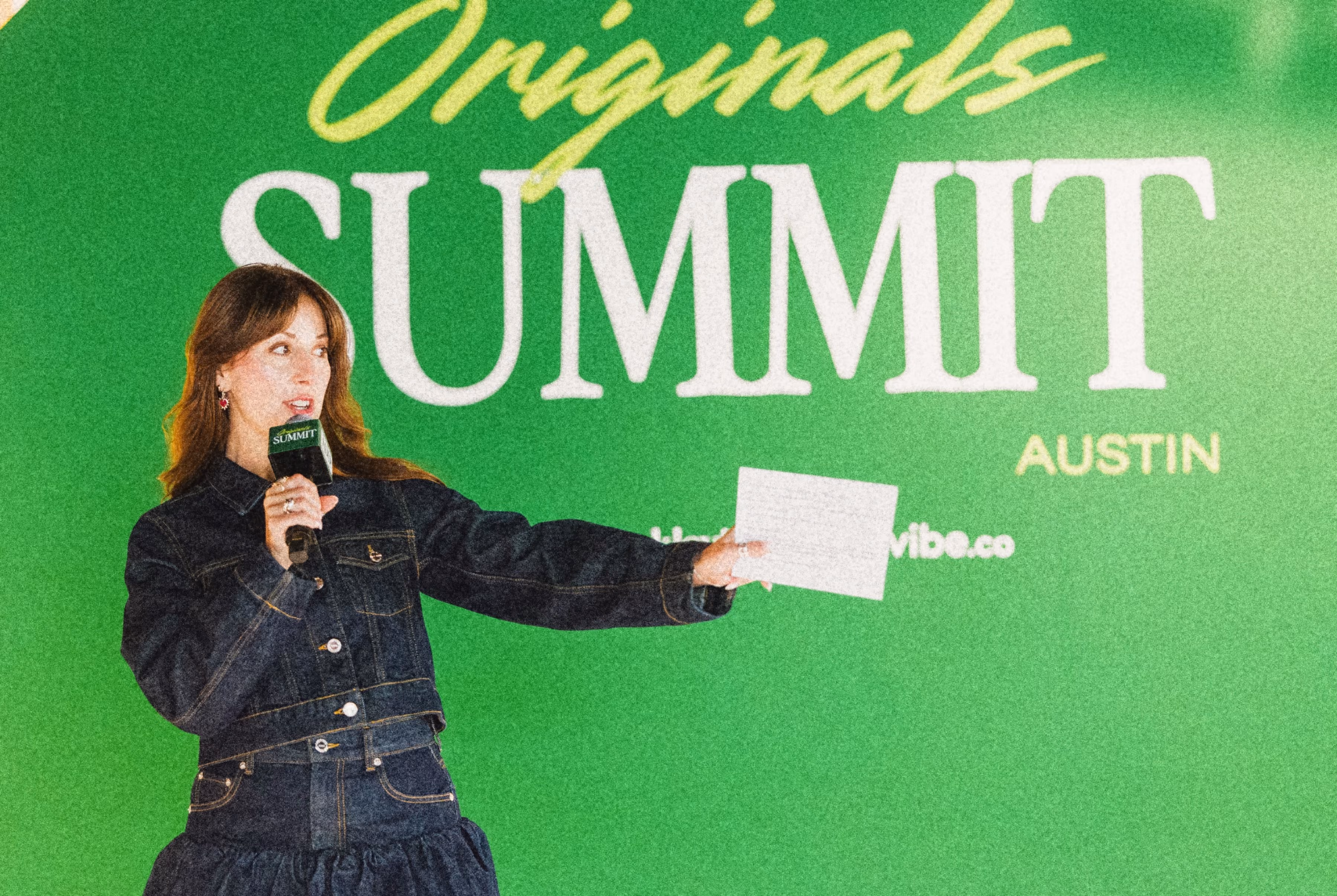 Woman speaking on stage holding microphone and notes in front of green Originals Summit Austin backdrop.