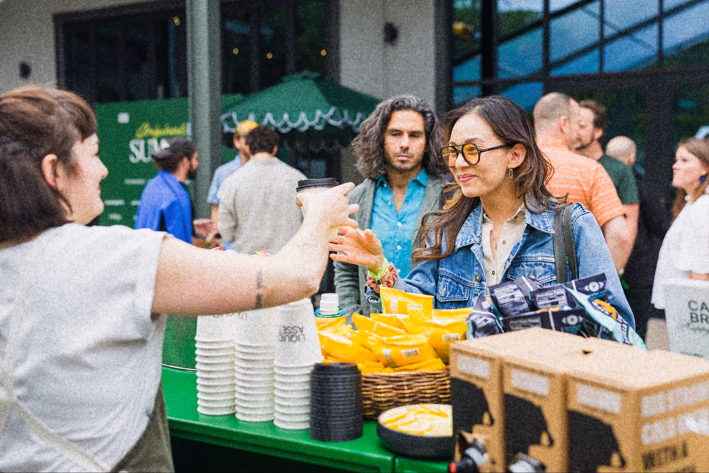 A woman in a denim jacket receives a cup from a vendor at an outdoor event with snacks displayed.