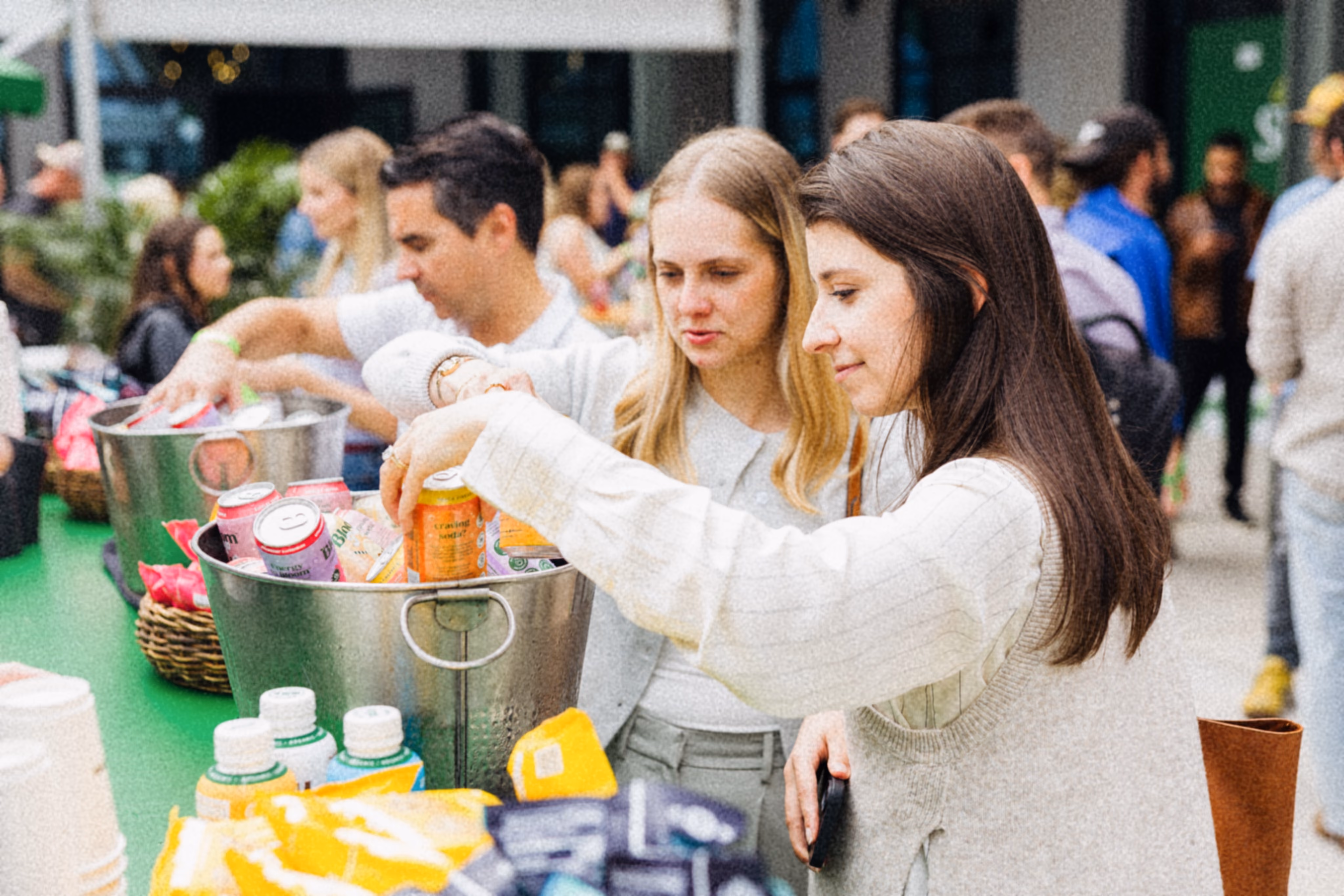 Three women engaged in conversation at an outdoor event, one holding a salad jar and another a pink drink can.