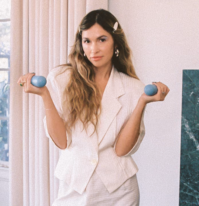 Woman in white suit holding small blue dumbbells with a neutral expression indoors.
