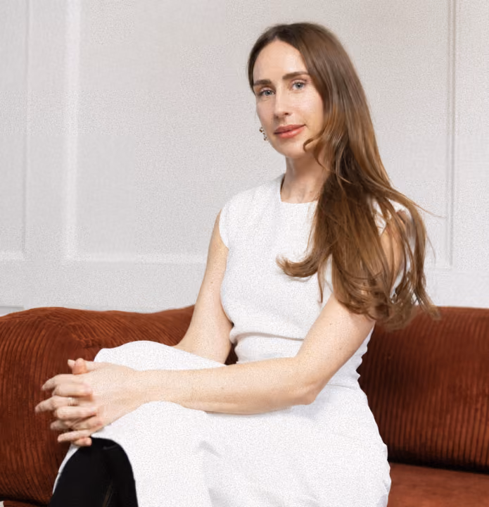 Woman with long brown hair wearing a sleeveless white top sitting on a red couch.