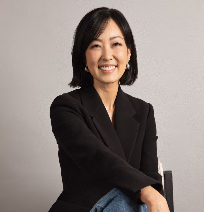 Smiling woman with black hair wearing a black blazer and sitting on a chair against gray background.