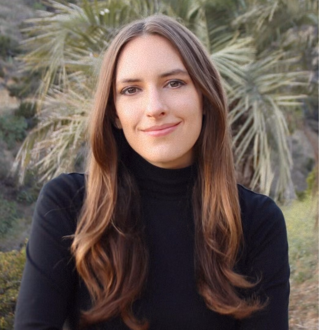 Smiling woman with long brown hair wearing black turtleneck, outdoors with palm trees in the background.