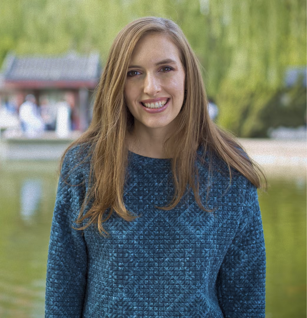 Smiling woman with long brown hair wearing black turtleneck, outdoors with palm trees in the background.