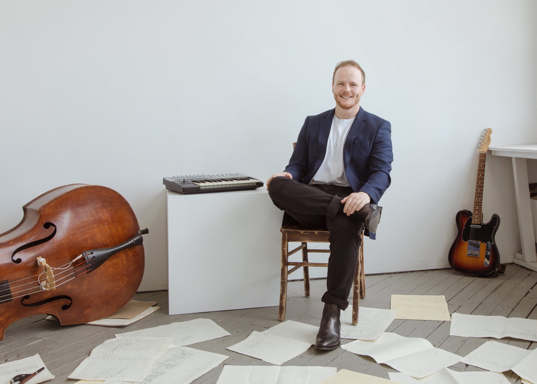 Composer Max Vinetz seated in a studio, an upright bass and electric guitar beside him and manuscript pages across the floor
