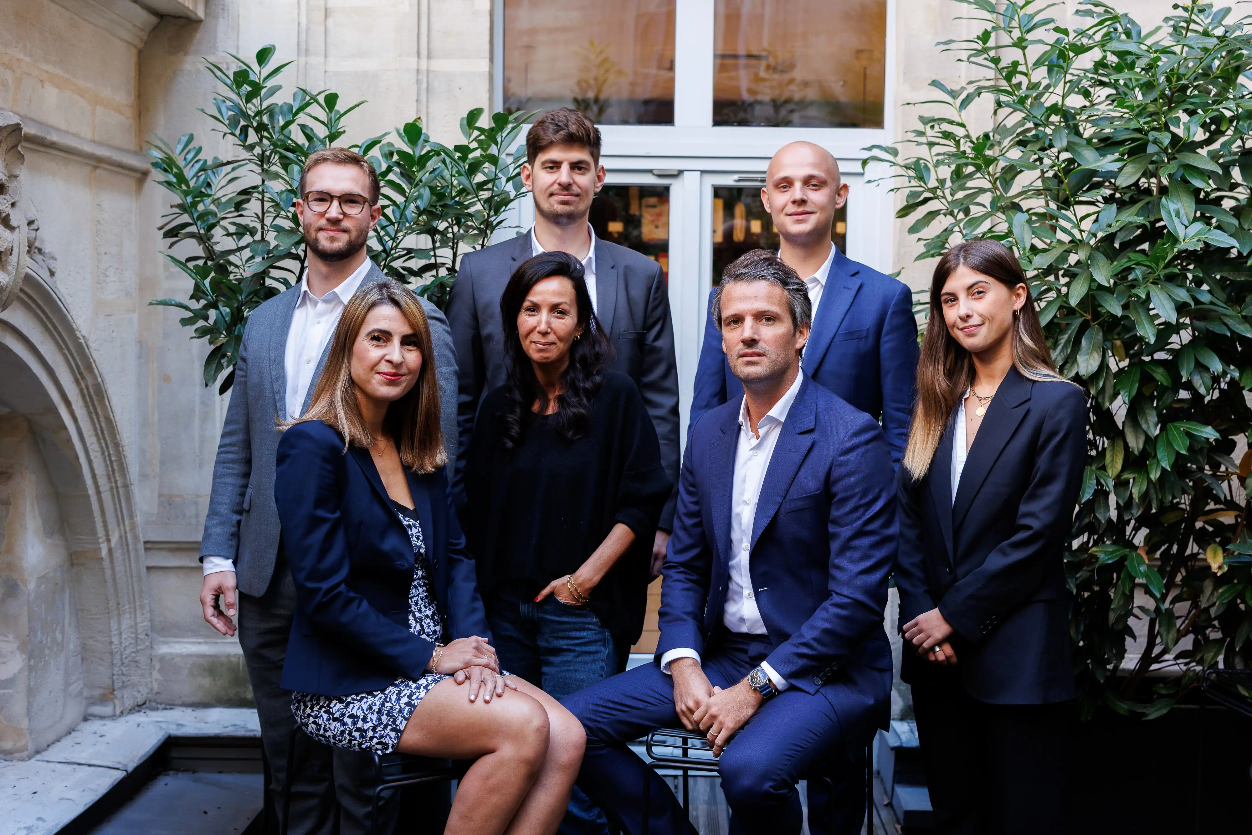 Group portrait of four men and three women in business attire, posed in front of a building entrance with greenery on each side.