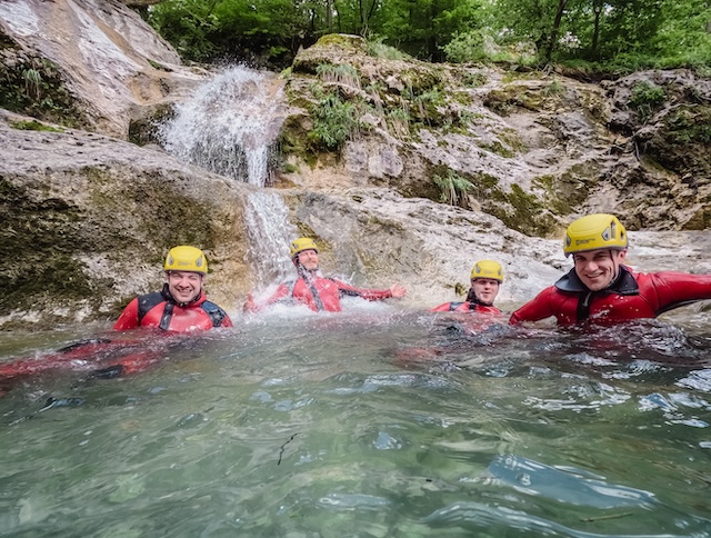 group of four friends sit in a clear blue pool below a small waterfall in a bled canyon