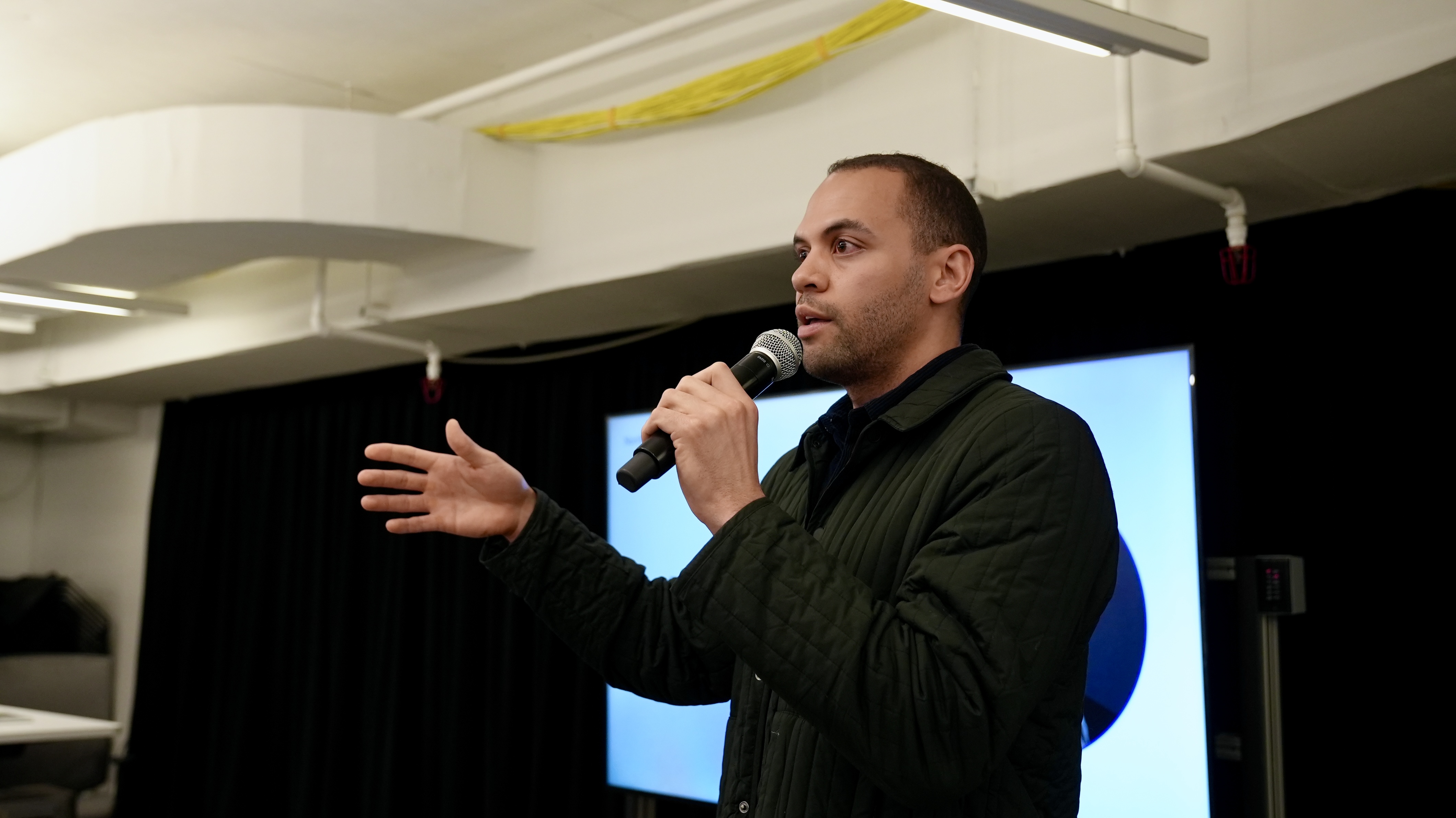 Payton stands at front of room in a black shirt holding a microphone