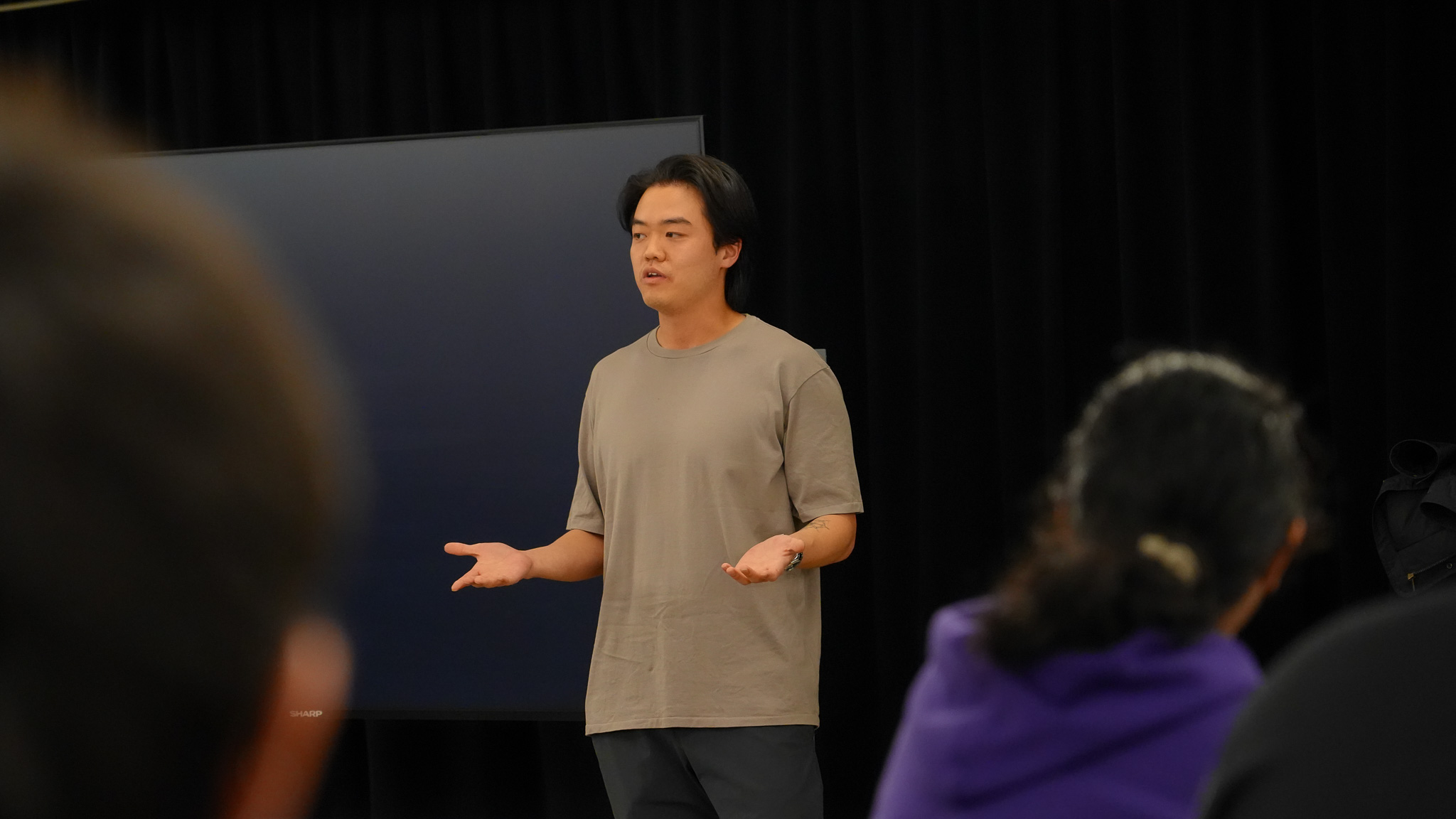 Kyle stands at the front of the room with his arms extended as he speaks