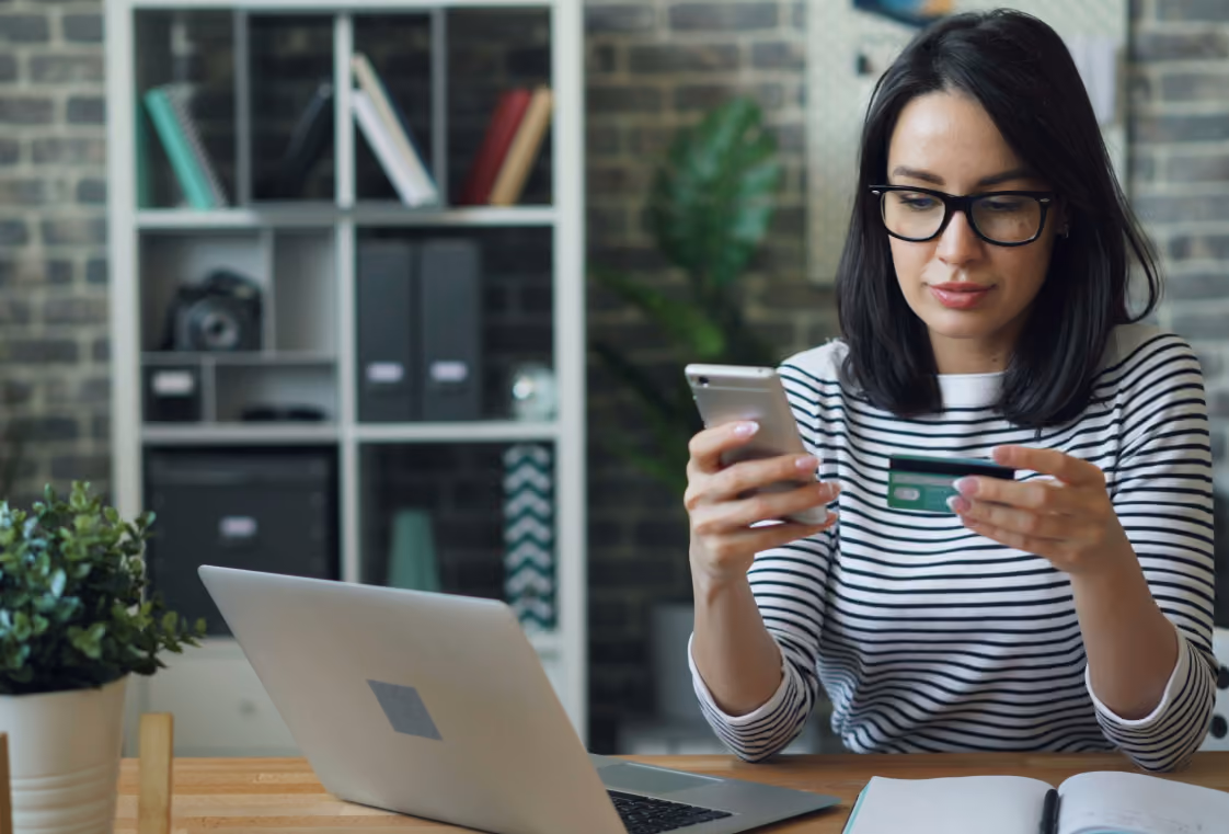 Woman at a wood desk using a smartphone and credit card beside a laptop in a modern office-style room