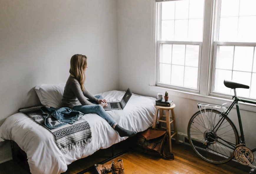 Woman typing on a laptop while sitting cross-legged on a white-dressed bed in a bright bedroom