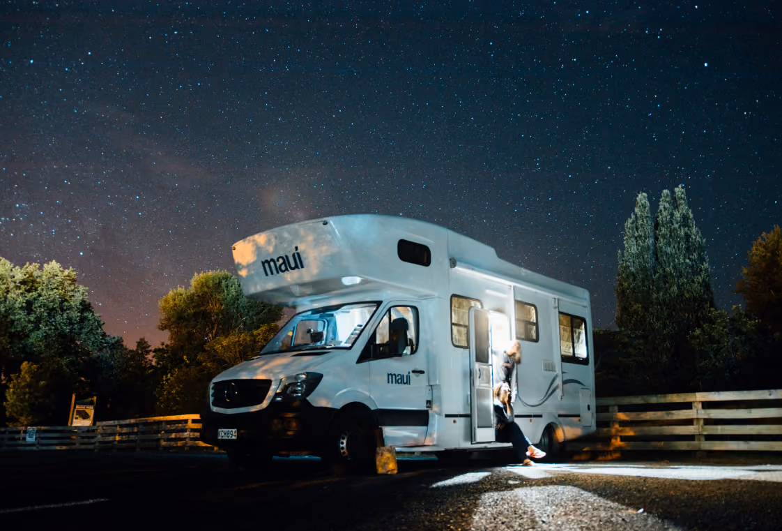 White Maui campervan parked at night under a starry sky with headlights illuminating the lot