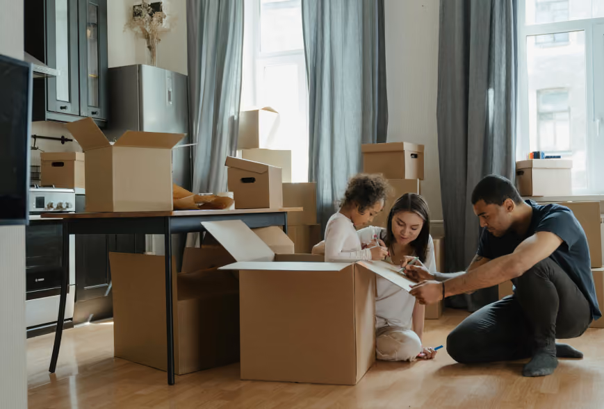 Family crouched on a wood floor decorating a moving box amid stacked cardboard boxes in a bright apartment