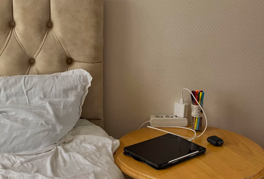 Round nightstand with a charging tablet, earbuds, power strip, and jar of markers beside a tufted bed