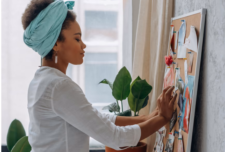 Woman in a teal headwrap pinning photos and notes to a cork mood board near a potted plant