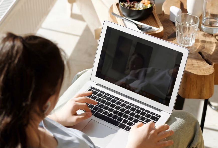 Over-the-shoulder view of someone on a laptop with earbuds beside a side table with a colorful meal bowl