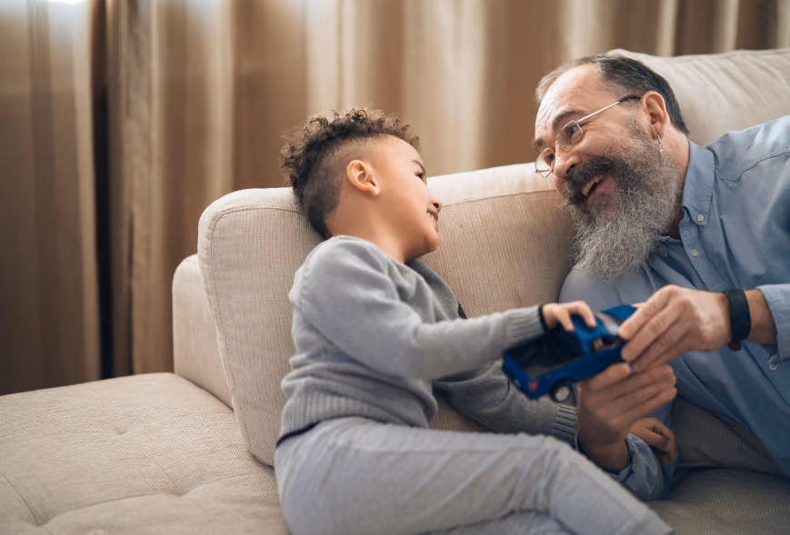 Smiling older man and young boy playing together with a blue toy truck on a beige sofa
