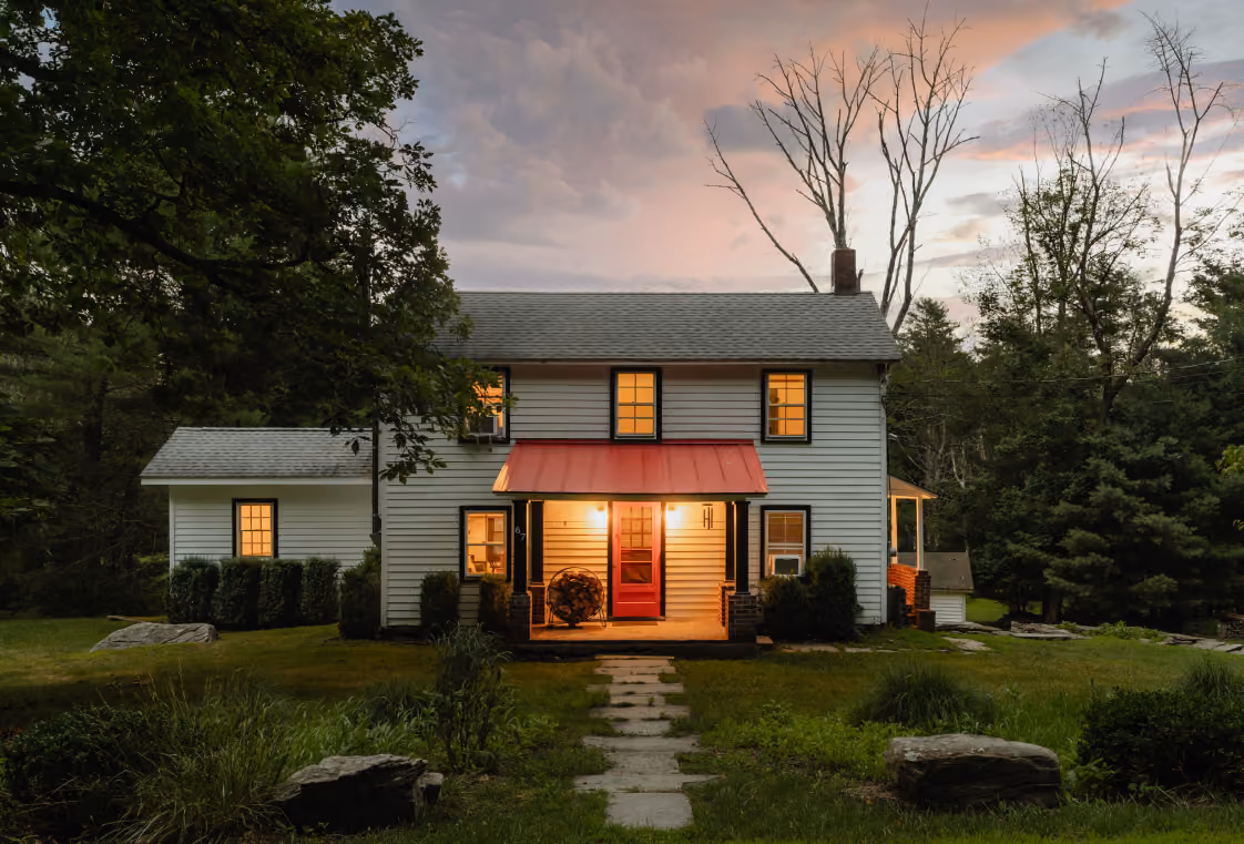 White farmhouse with black trim, red porch roof, and glowing windows under a pink sunset sky