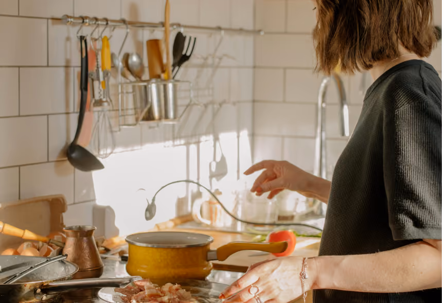Person cooking at a stove in a sunlit kitchen with subway tile, hanging utensils, and eggs on the counter