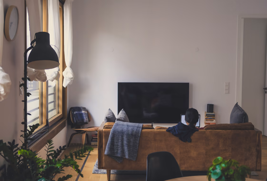 Back view of someone on a sofa using a laptop toward a wall TV in a bright plant-filled living room
