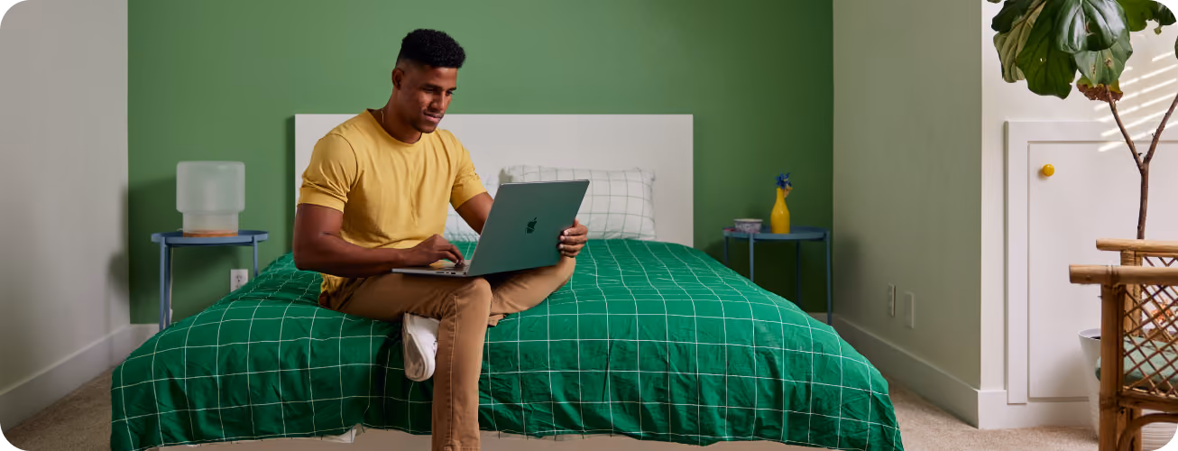 Person on a bed with a green grid duvet using a laptop in a bedroom with dark green accent wall