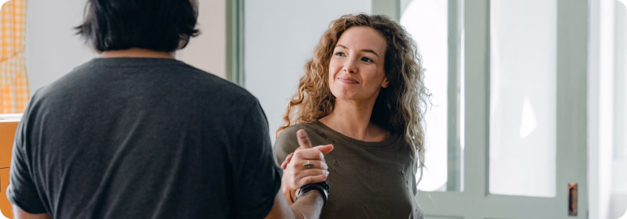 Smiling woman with curly blonde hair holds hands with a partner seen from behind in a bright doorway
