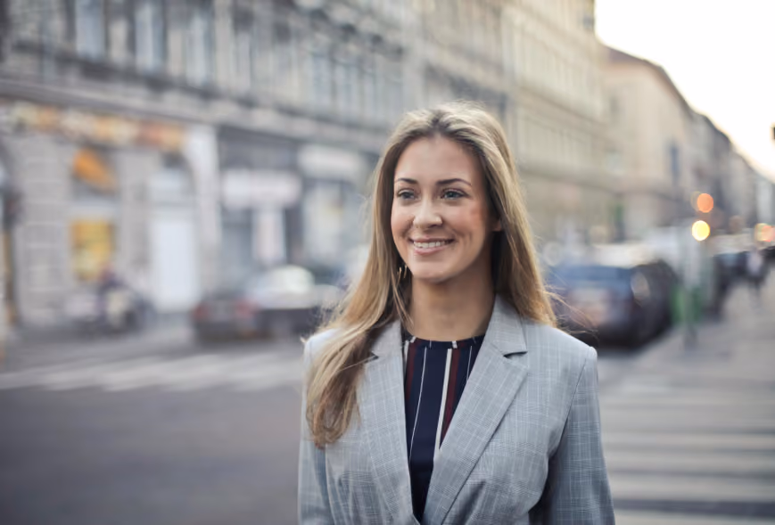 Professional young woman in a grey blazer smiling at a sunny urban crosswalk
