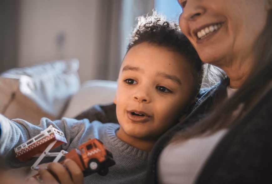 Young boy with a toy fire truck beside a smiling older woman on a cozy sofa