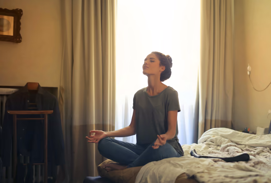 Woman meditating cross-legged on a bed in front of a bright window with soft natural light
