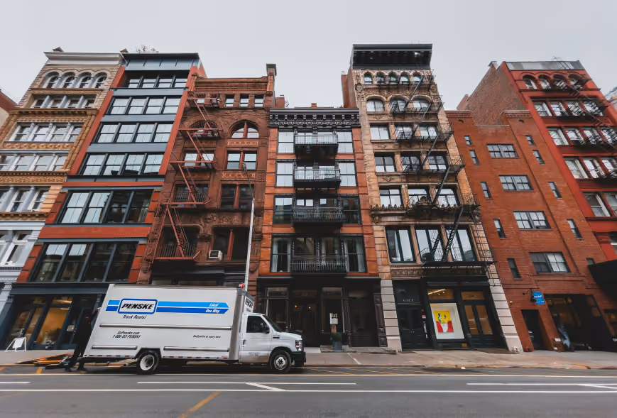 Penske moving truck parked along a row of brick and stone city apartment buildings