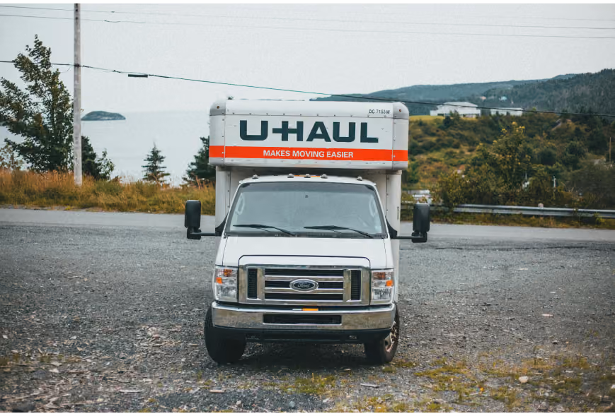 Front view of a white U-Haul truck parked on gravel near water and green hills