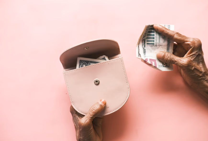 Older hands opening a pink coin purse beside a crumpled five-dollar bill on a solid pink surface