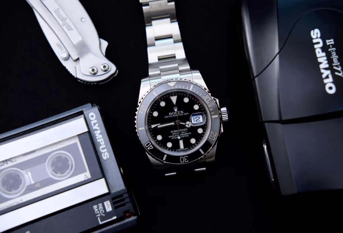 Flat lay of a stainless dive watch with vintage recorder, compact film camera, and folding knife on black