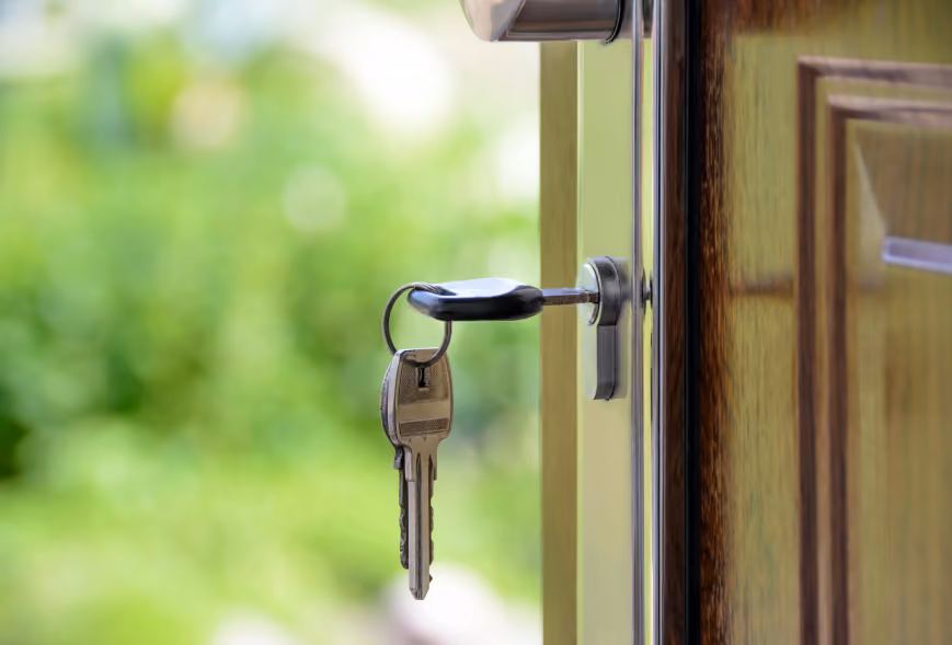 Keys inserted in the lock of a slightly open wood door with a blurred green garden beyond