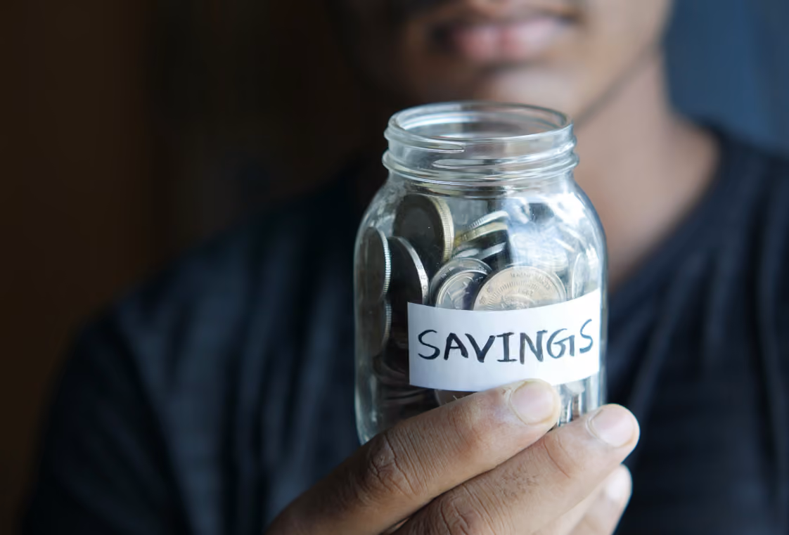 Hand holding a glass jar of coins labeled SAVINGS in bold handwriting