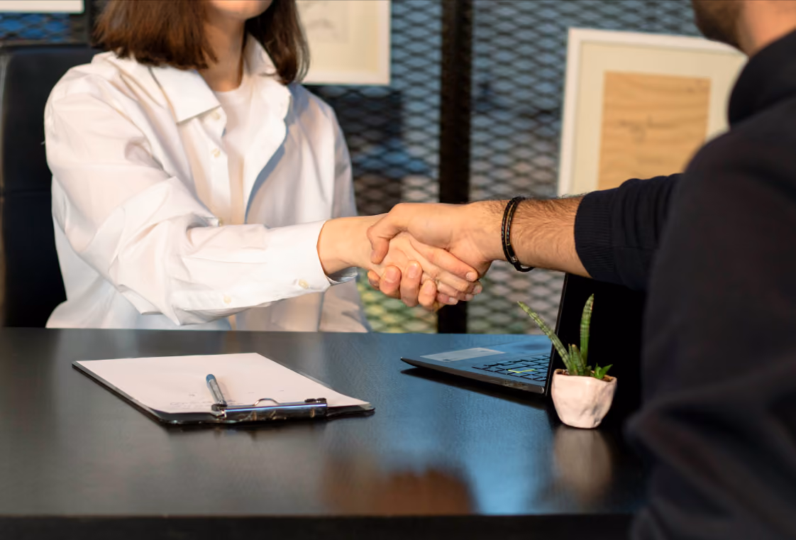 Handshake across a black desk with clipboard, laptop, and small potted plant in a modern office