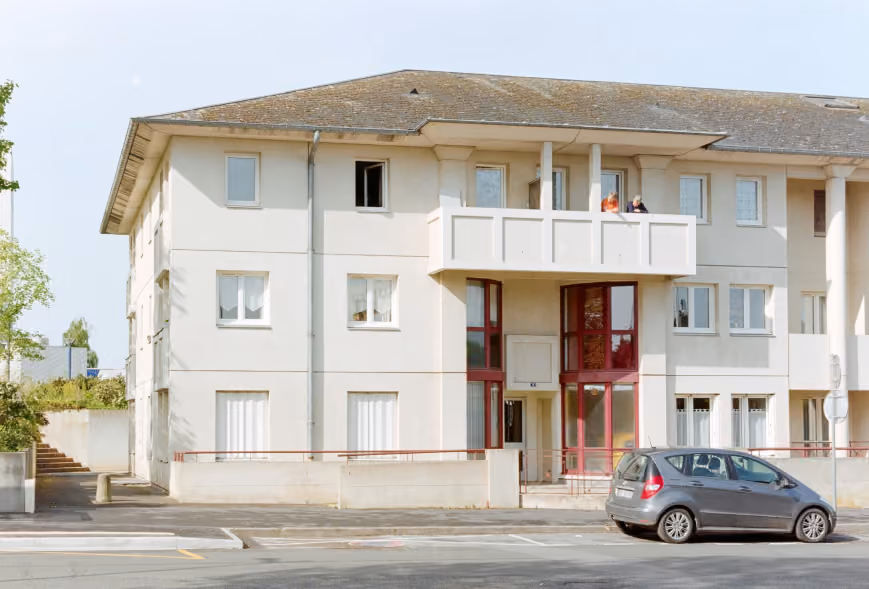 Three-story cream apartment building with people on a balcony above a maroon-trimmed entrance