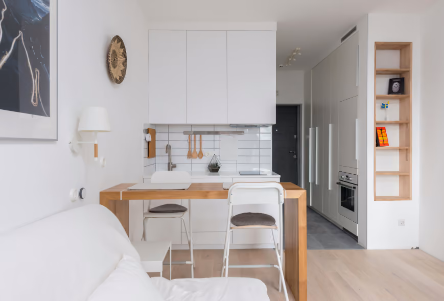 Bright studio apartment with white kitchen cabinets, wood dining table, and floor-to-ceiling wardrobes