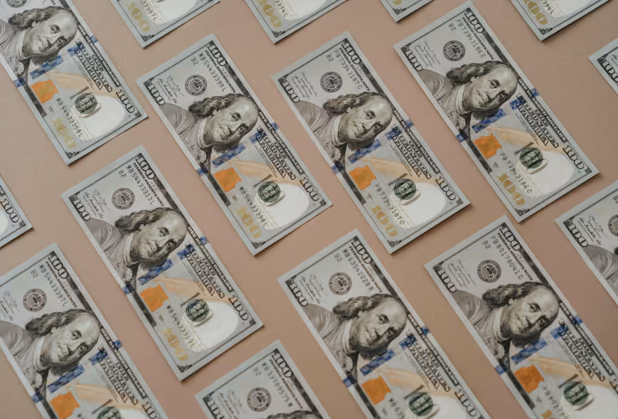 Overhead grid of US hundred-dollar bills spaced neatly on a tan background