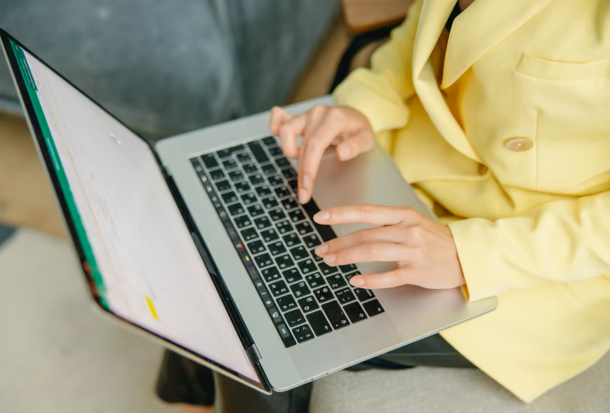 Close-up of hands typing on a laptop balanced on the lap of someone in a yellow blazer