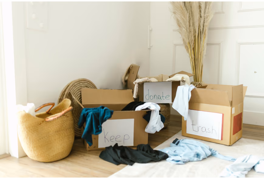 Three sorting boxes labeled keep, donate, and trash with clothes on a hardwood floor