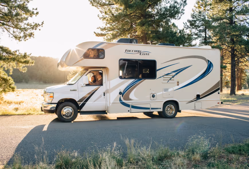 Smiling couple visible in the cab of a Class C motorhome parked along a pine forest road