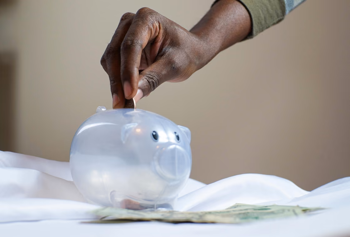 Hand dropping a coin into a translucent piggy bank above scattered dollar bills